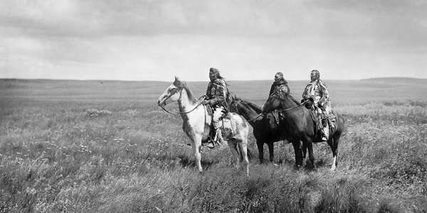 black and white photo of three indigenous men sitting on horses in the grass prairies