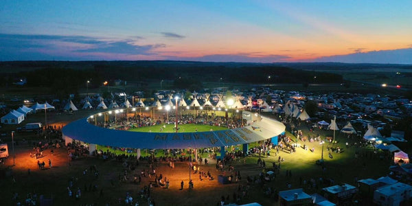 aerial view at dusk with a round arena with open air in the middle and teepees surrounding the arena with lots of people in and around the arena