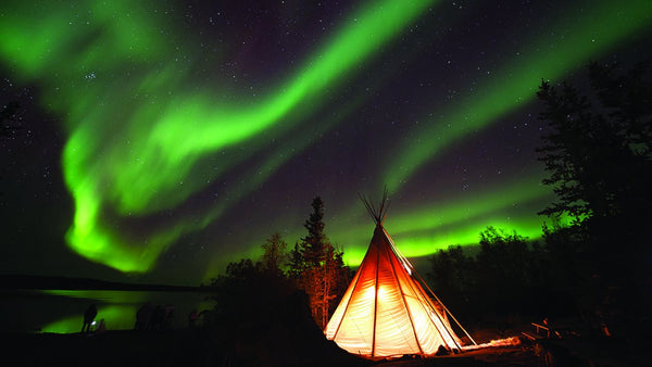 nighttime with a teepee in the foreground lit from the inside and green northern lights in the sky
